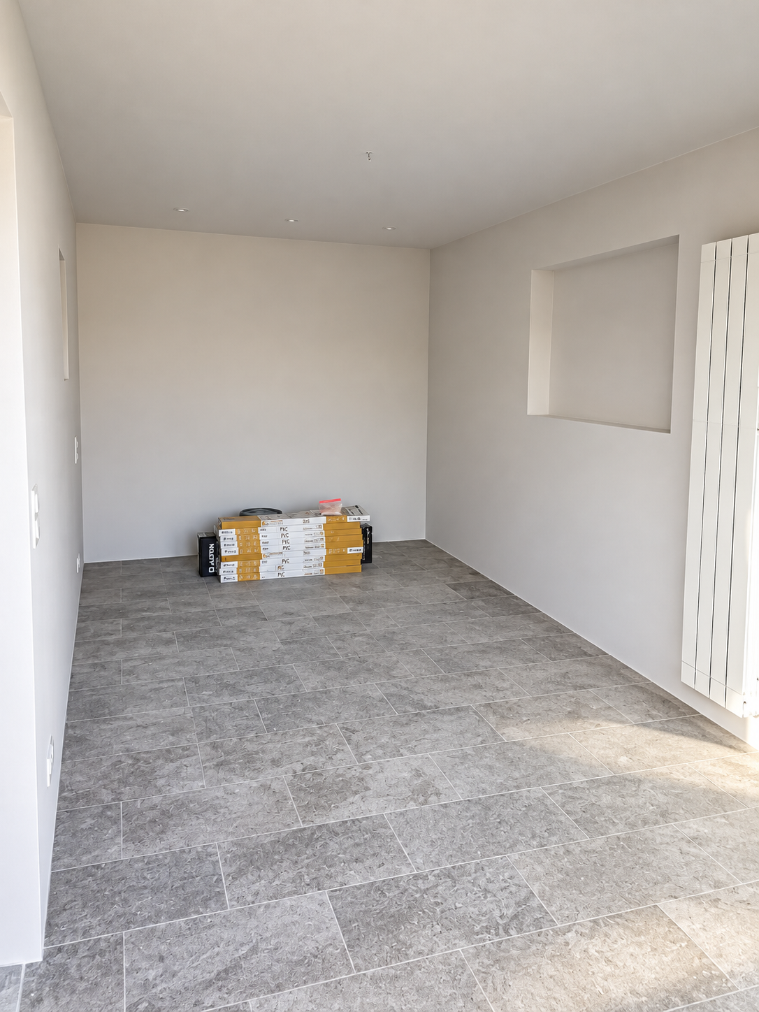 Pièce aux murs clairs, sol gris à grands carreaux, niche rectangulaire et radiateur vertical, avec des paquets au fond.
