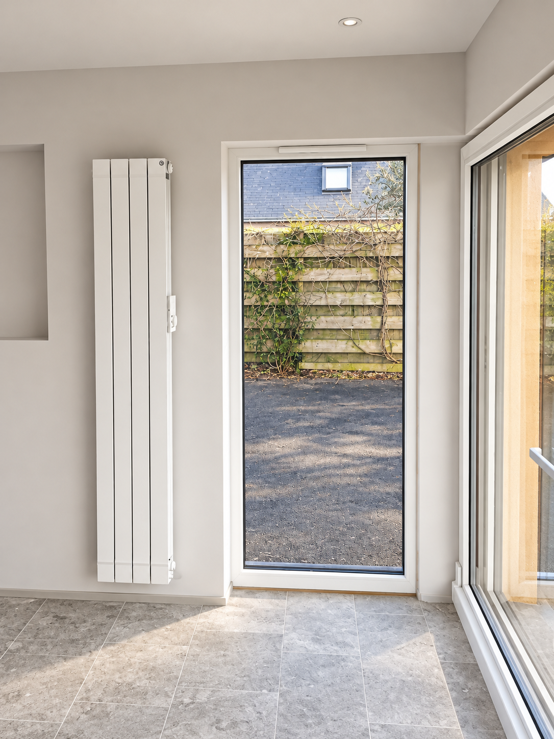 Vitrage étroit près d’une grande baie vitrée, radiateur vertical blanc et sol gris.